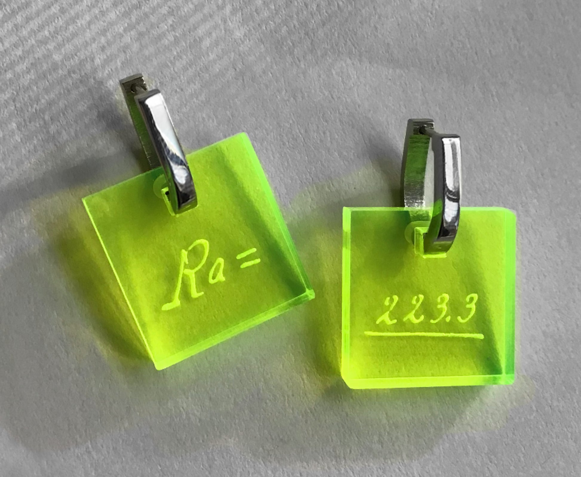 Neon acrylic earrings engraved with Marie Curie’s handwriting and the atomic weight of radium, stainless steel leverbacks, looking from above.