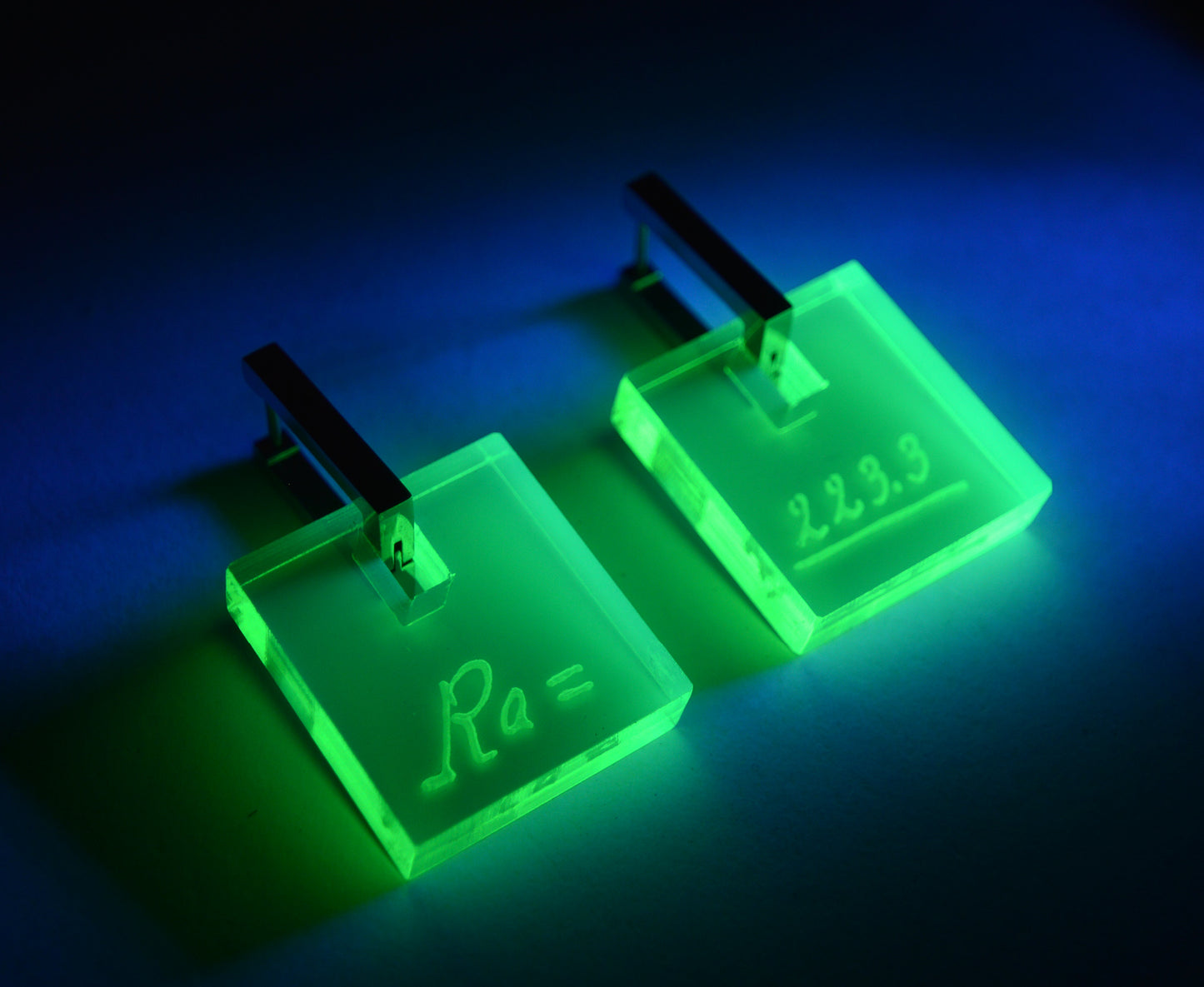 Neon acrylic earrings engraved with Marie Curie’s handwriting and the atomic weight of radium, glowing under blacklight with stainless steel leverbacks.
