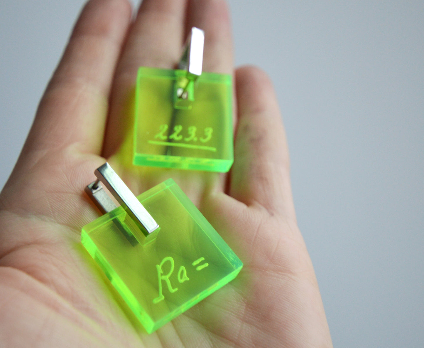 Neon acrylic earrings engraved with Marie Curie’s handwriting and the atomic weight of radium, in palm of hands, with stainless steel leverbacks.